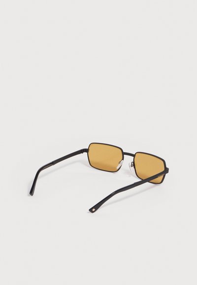 Lunettes de soleil à monture rectangulaire noire avec des verres teintés jaunes reposant sur une surface blanche plate.