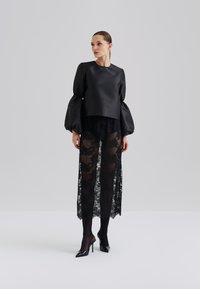 Svart sateng topp med ballongsleeves lagd over gjennomsiktige svarte blonderbukser. Ensemblet har svarte høye spisse sko.