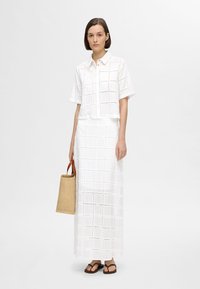 Hvid bomulds crop-top med firkantede øjeletmønstre, der er parret med en lang hvid nederdel med lignende mønstre. Modellen holder en beige vævet taske.