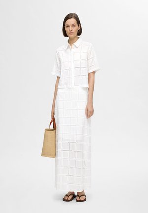 Chemise blanche en coton courte avec des motifs en œillets carrés, associée à une longue jupe blanche avec des motifs similaires. Le mannequin tient un sac en osier beige.