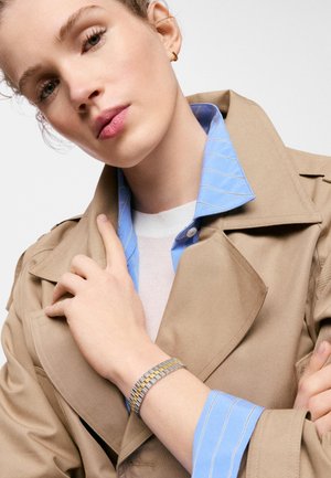 Joven mujer de piel clara y ojos verdes lleva un abrigo tipo trench beige sobre una camisa azul a rayas, complementado con una pulsera de oro y plata y un pequeño aro.