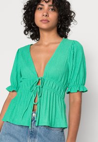 Blusa verde texturada com decote em V profundo, mangas bufantes curtas e fecho de laço na frente. Combinada com denim azul.