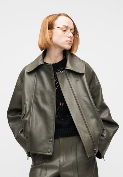 Νέος με κοντά κόκκινα μαλλιά και γυαλιά που φοράει δερμάτινο μπουφάν και παντελόνι ελιάς, μαύρο πλεκτό τοπ, τα χέρια στις τσέπες, κοιτάζει αριστερά.