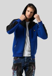 Blau- und schwarze Zip-Hoodie mit Kapuze, ausgestattet mit kontrastierenden Einsätzen, Seitentaschen und einem Logo auf der Brust. Glatte Textur.