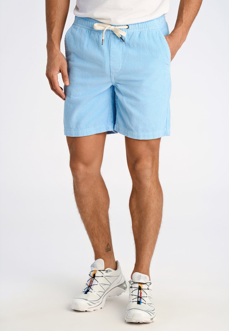  bañador de rayas azul claro con cinturilla ajustable, bolsillos laterales y un corte relajado, combinado con zapatillas deportivas blancas.