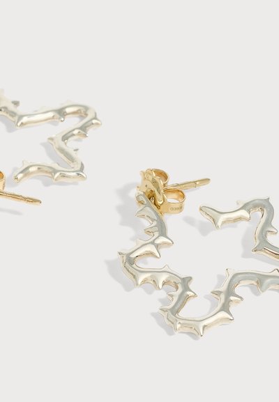 Boucles d’oreilles créoles en argent avec des bords irréguliers et pointus et des attaches clous couleur or sur un fond clair.