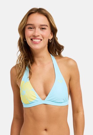 Młoda kobieta uśmiecha się, nosząc jasnoniebieski top bikini na szyję z dużym żółtym kwiatem po lewej stronie.