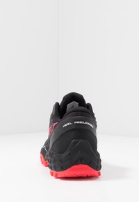 Černá sportovní bota s texturovaným svrškem, výraznou oporou paty označenou "HEEL PRELOADER" a červenou podešví s robustním vzorem trakce.