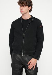 Melna džinsa jaka ar rāvējslēdzēja aizdari, sprādzes apkakli un smalku sarkanu logotipa detaļu. Raksturota ar pieguļošu siluetu un teksturētu audumu.