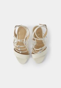 Sandales à talons blancs avec une texture lisse, dotées de brides de cheville et d'accents en cristal décoratifs, à bout fermé et à talon modeste.