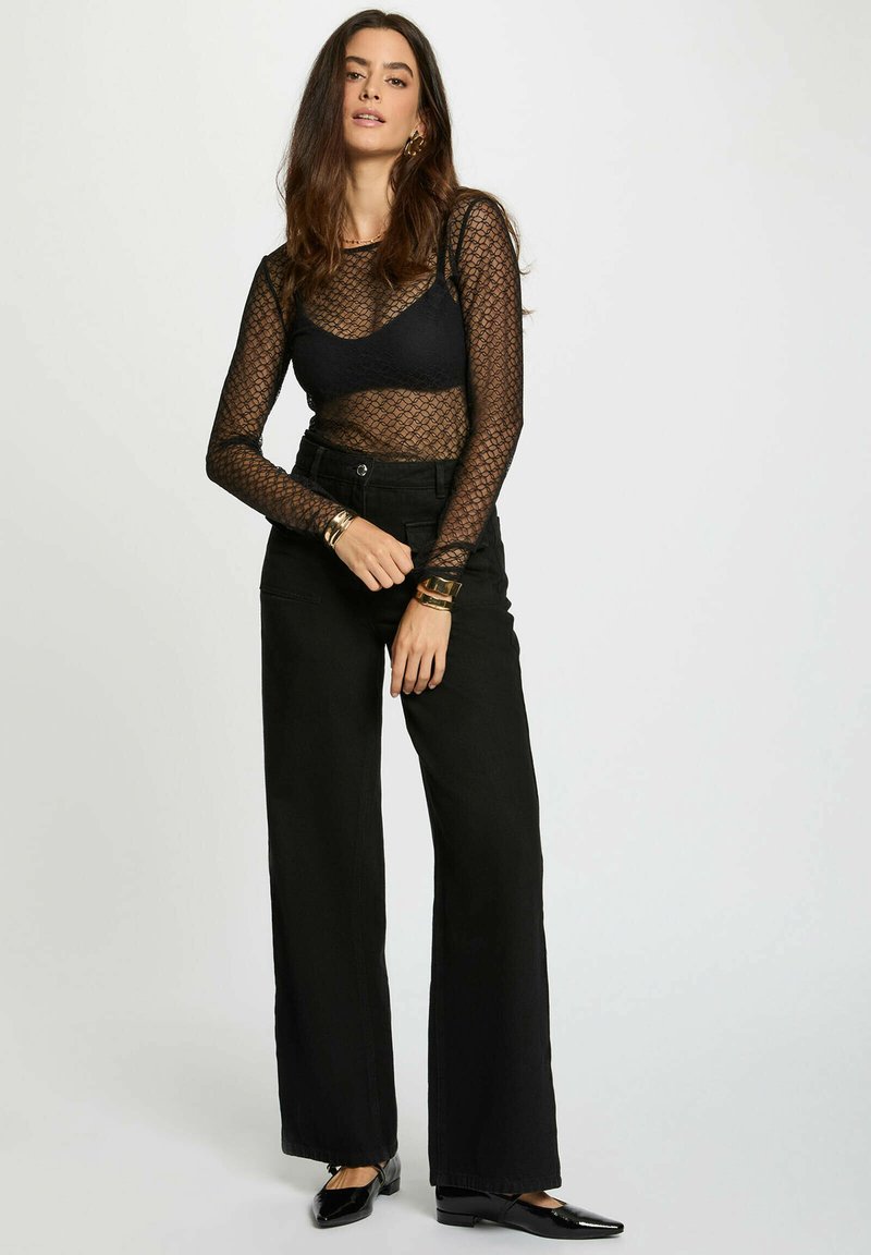 Donna in piedi con una mano che stringe l'altro polso, indossando un top a maniche lunghe in rete nera sopra un bralette nero, pantaloni a vita alta e scarpe nere.