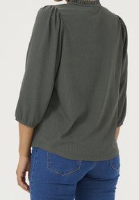 Blusa verde scuro con texture, collo arruffato e maniche a sbuffo, abbinata a jeans in denim blu. Design minimalista e vestibilità morbida.