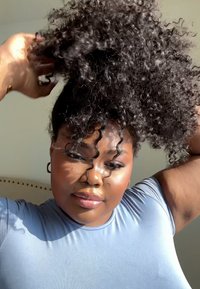 Svart krøllete hår blir stylet til en hestehale. Personen har på seg en tettsittende lyseblå topp med en glatt tekstur og en naturlig glød.