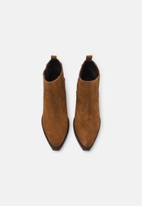 Bottes chevilles en suède marron avec unempot pointu, dotées d'un talon bas et de languettes sur les côtés. Texture lisse avec des détails de couture minimaux.
