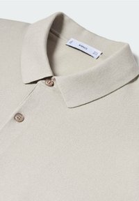 Beige polo-paita, jossa on teksturoitu materiaali, klassinen kaulus ja kaksi nappia edessä. Tunnusnäyttöön on merkitty brändilogo "MNG."