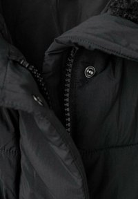 Primer plano de una chaqueta de invierno negra que muestra el cierre, el botón a presión y parte de la capucha forrada de pelo.