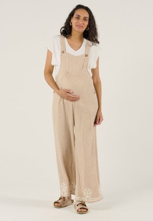 Zwangere vrouw draagt beige overalls met witte bloemenborduursel, wit T-shirt en beige sandalen, houdt voorzichtig haar buik vast.