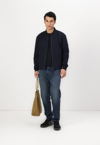 Giacca bomber navy, chiusura con zip, maniche a costine; t-shirt nera, jeans blu, sneakers nere. Borsa a tracolla beige con spallacci.