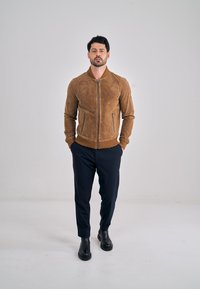 Ruskea mokkanahkainen bomber-takki vetoketjulla, resorikahvoilla ja kahdella sivutaskulla, yhdistetty tummiin housuihin ja mustiin nilkkureihin.