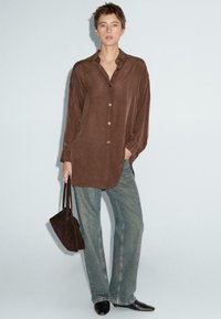 Camicia marrone a maniche lunghe con bottoni e una texture liscia, abbinata a jeans larghi blu chiaro e una borsa marrone scuro. Scarpe nere a punta.