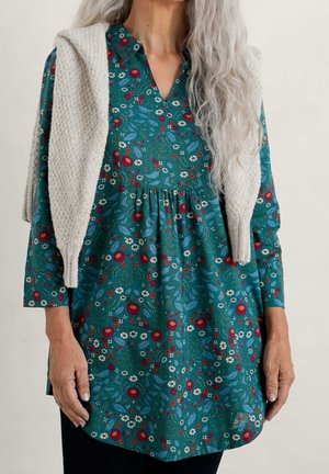 Vrouw met lang grijs haar draagt een groene bloemen tuniek en een beige gebreide trui over haar schouders gedrapeerd, staand tegen een effen achtergrond.