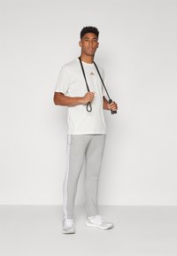 Lys grå træningsbukser med hvide sidelister og en lyse farvet T-shirt med rund hals. Modellen holder et sort sjippetov.