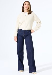 Hellbeige, flauschiger Strickpullover mit langen Ärmeln, kombiniert mit hochtaillierten, dunkelblauen Schlagjeans und metallischen, spitz zulaufenden Schuhen.