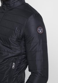 Schwarze Steppjacke mit genoppter Textur, hohem Kragen, Reißverschlusstaschen und einem runden Logo-Patch am linken Ärmel. Glattes, glänzendes Material.