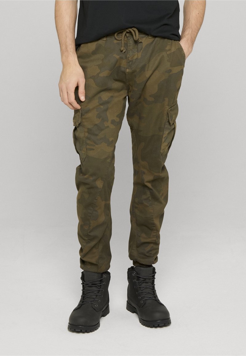 Homme portant un pantalon cargo camouflage vert olive et des bottes noires à lacets, debout sur un fond uni gris clair.