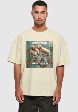 Beige oversized cotton T-shirt featuring a floral graphic with cotton bolls and the text “COTTON CLUB” and “2Y STUDIOS.” Text in white and blue.