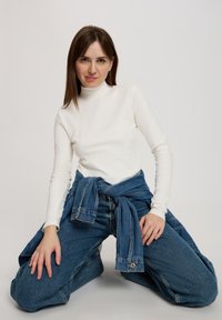 Biała, długiego rękawa bluzka z golfem w połączeniu z luźnymi niebieskimi dżinsami, związanym w talii. Modelka klęczy i pozuje na jednolitym tle.