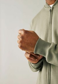 Chaqueta de cremallera verde claro con puños acanalados, mostrada en un primer plano sobre una mano con piel texturada; se puede ver el detalle de la tela y el ajuste.