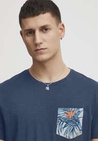 Laivansinis puuvillapaita, jossa on neliön muotoinen tasku, jonka koristeena on trooppinen kukka- ja lehtikuosi; hopeaketju, jossa on charmikiinnike, jossa on kiinalaisia merkkejä.