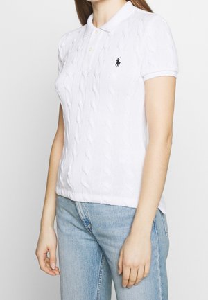 Femme portant un polo blanc à manches courtes en maille torsadée avec un logo noir brodé, associé à un jean bleu clair.
