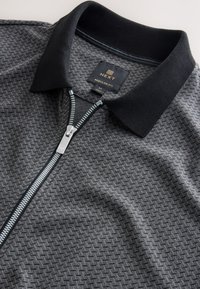 Grå mønstrede zip-up skjorte med sort krave, sølv lynlås og "NEXT Regular Fit" label indeni kraven, der viser størrelse medium.