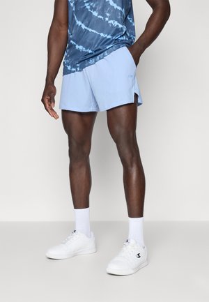 Hellblaue Sportshorts mit seitlichen Netzpaneelen, kombiniert mit einem blauen Batik-Top. Weiße Sneakers und Socken runden das Outfit ab.