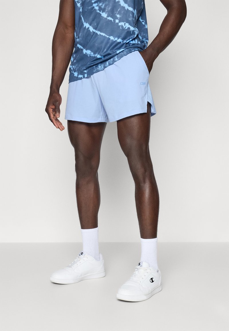 Lyseblå træningsshorts med sidepaneler i mesh, parret med en blå tie-dye tanktop. Hvide sneakers og sokker fuldender outfittet.