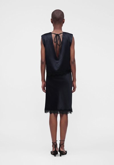 Personne aux cheveux courts portant une robe noire en satin sans manches avec un dos en V profond noué au cou et des sandales à talons noires, debout sur fond blanc.