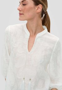 Weiße Bluse mit einem strukturierten Blumenmuster, V-Ausschnitt, Drei-Viertel-Ärmeln und Quastenverzierung am Ausschnitt.