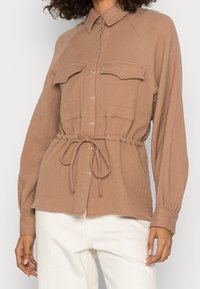 Bruine textuurblouse met knoopsluiting, twee voorzakken en een cinched taille. Lange mouwen met knoopsluiting bij de manchet. Gestyled over witte broek.