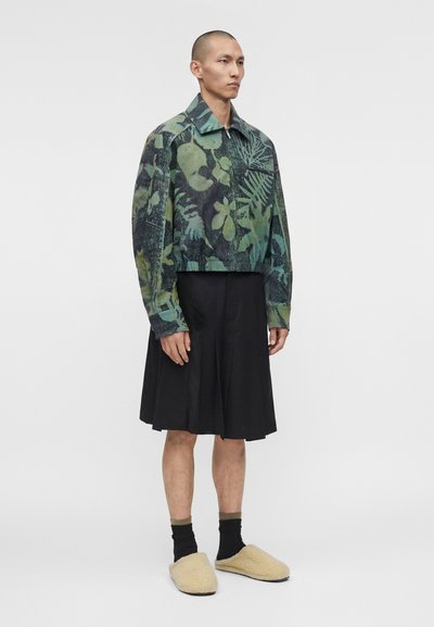 Άνδρας ντυμένος με πράσινο και μαύρο σακάκι με φλοράλ μοτίβο, μαύρη πλισέ φούστα, μαύρες κάλτσες και μπεζ χνουδωτές παντόφλες, στέκεται μπροστά σε λευκό φόντο.