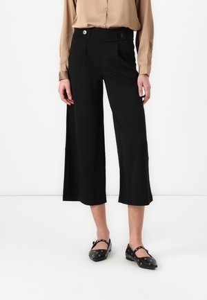 Femme portant un pantalon noir large et court taille haute avec des boutons à la taille, un chemisier beige à manches longues et des ballerines Mary Jane noires avec œillets.