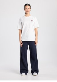 Wit katoenen T-shirt met korte mouwen en een klein bedrukt ontwerp op de linkerborst, gecombineerd met marineblauwe wijde broeken en witte schoenen.