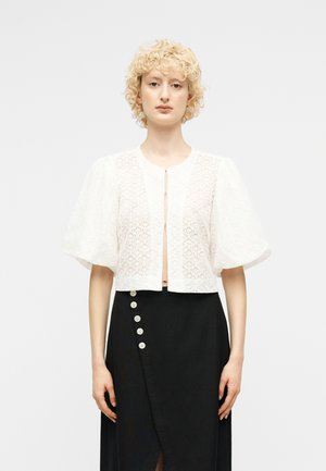 Ξανθιά γυναίκα με κατσαρά μαλλιά φορεί μια λευκή δαντελένια κοντή μπλούζα με φουσκωτά μανίκια και μια μαύρη φούστα με ψηλή μέση και πλευρικά κουμπιά.