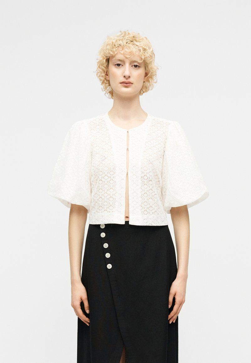 Ξανθιά γυναίκα με κατσαρά μαλλιά φορεί μια λευκή δαντελένια κοντή μπλούζα με φουσκωτά μανίκια και μια μαύρη φούστα με ψηλή μέση και πλευρικά κουμπιά.