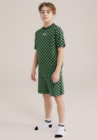 Zeleni karirani komplet s t-shirtom in kratkimi hlačami, črnim ovratnikom. Tiskani vzorec vključuje majhne črne akcentne detajle. Nosite z belimi nogavicami.