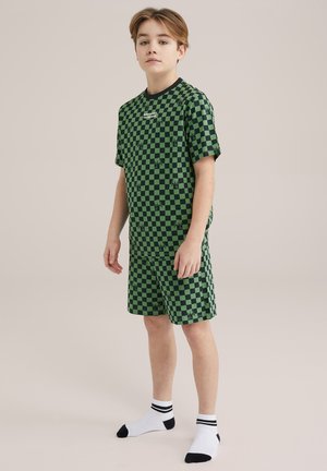 Grünes karierter T-Shirt- und Shorts-Set mit einem schwarzen Ausschnitt. Das Druckmuster enthält kleine schwarze Akzente. Getragen mit weißen Socken.