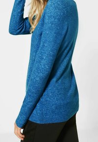 Kvinna som bär en långärmad blå stickad tröja med svarta byxor, står i profil mot en vit bakgrund.