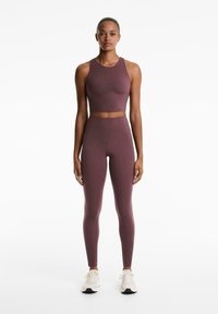 Egyenes álló nő, aki egymáshoz illő lila crop topot és magas derekú leggingst visel, fehér sportcipővel egy sima fehér háttér előtt.