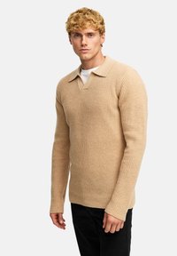 Beige strikket genser med V-hals og krage, med et teksturert mønster. Modellen har på seg svarte bukser og står foran en ensfarget bakgrunn.